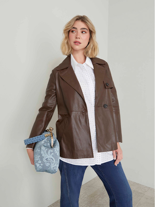 Modella indossa giacca in similpelle di colore marrone abbinata a camicia di colore bianco e jeans dal lavaggio blu scuro.